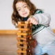 Kleines Mädchen spielt mit einem Turm aus Holzklötzen und legt einen Klotz auf die Spitze, neutraler Hintergrund.