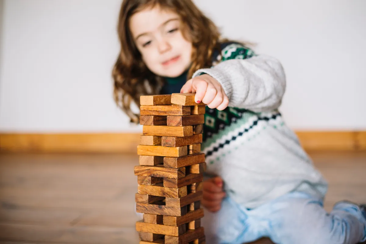 Kleines Mädchen spielt mit einem Turm aus Holzklötzen und legt einen Klotz auf die Spitze, neutraler Hintergrund.