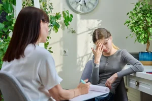 Zwei Personen in einer Neurorehabilitationssitzung: Eine Frau macht sich Notizen, während eine junge Frau in therapeutischer Umgebung besorgt wirkt.