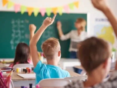 Kindergruppe im Klassenraum bei aufmerksamem und planendem Arbeiten, während eine Lehrerin an die Tafel schreibt