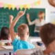 Kindergruppe im Klassenraum bei aufmerksamem und planendem Arbeiten, während eine Lehrerin an die Tafel schreibt