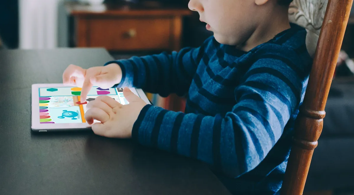 Kleines Kind sitzt an einem dunklen Tisch und nutzt ein Tablet, um Übungen mit NeuronUP2GO zu bearbeiten; auf dem Bildschirm sind Farben und Grafiken zu sehen.