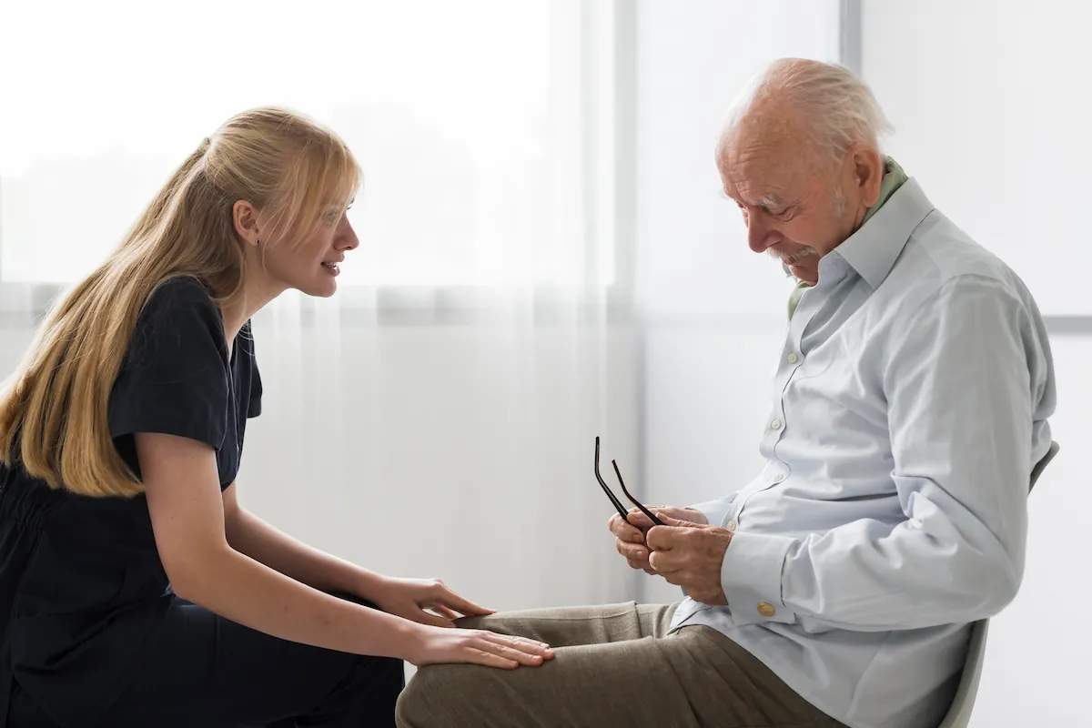 Junge Pflegeperson unterhält sich in einer hellen Umgebung mit einer älteren Person mit Demenz und unterstützt die Interaktion durch Gesten.