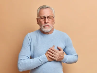 Älterer Mann mit grauem Haar und Bart, Brille und hellblauem Pullover, der sich mit beiden Händen an die Brust fasst, vor beigem Hintergrund.