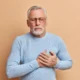 Älterer Mann mit grauem Haar und Bart, Brille und hellblauem Pullover, der sich mit beiden Händen an die Brust fasst, vor beigem Hintergrund.
