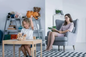 Kind sitzt mit Spielzeug an einem Tisch und wird in einer hellen Lernumgebung von einer erwachsenen Frau beobachtet.