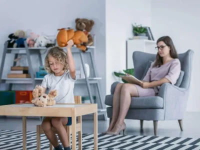 Kind sitzt mit Spielzeug an einem Tisch und wird in einer hellen Lernumgebung von einer erwachsenen Frau beobachtet.