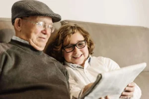 Grand-père portant une casquette et des lunettes, assis sur un canapé à côté de son petit-fils, tous les deux regardant un journal et partageant un moment d’apprentissage.
