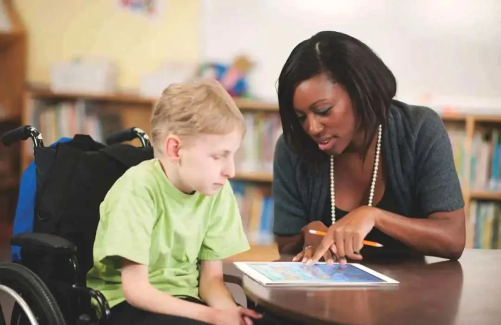 Une ergothérapeute enseigne à un enfant en fauteuil roulant à l'aide d'une tablette, dans un environnement scolaire.