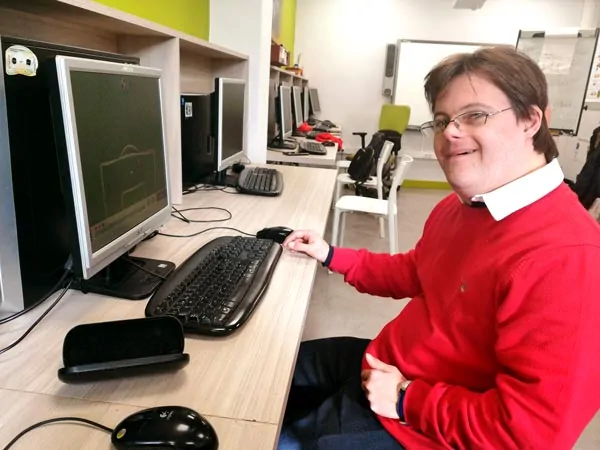 Personne atteinte du syndrome de Down souriante devant un ordinateur dans un centre occupationnel, avec des équipements informatiques en arrière-plan.