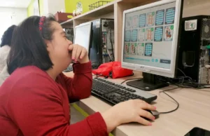 Une fille aux cheveux foncés assise devant un ordinateur dans une salle de classe colorée, concentrée sur une activité éducative.