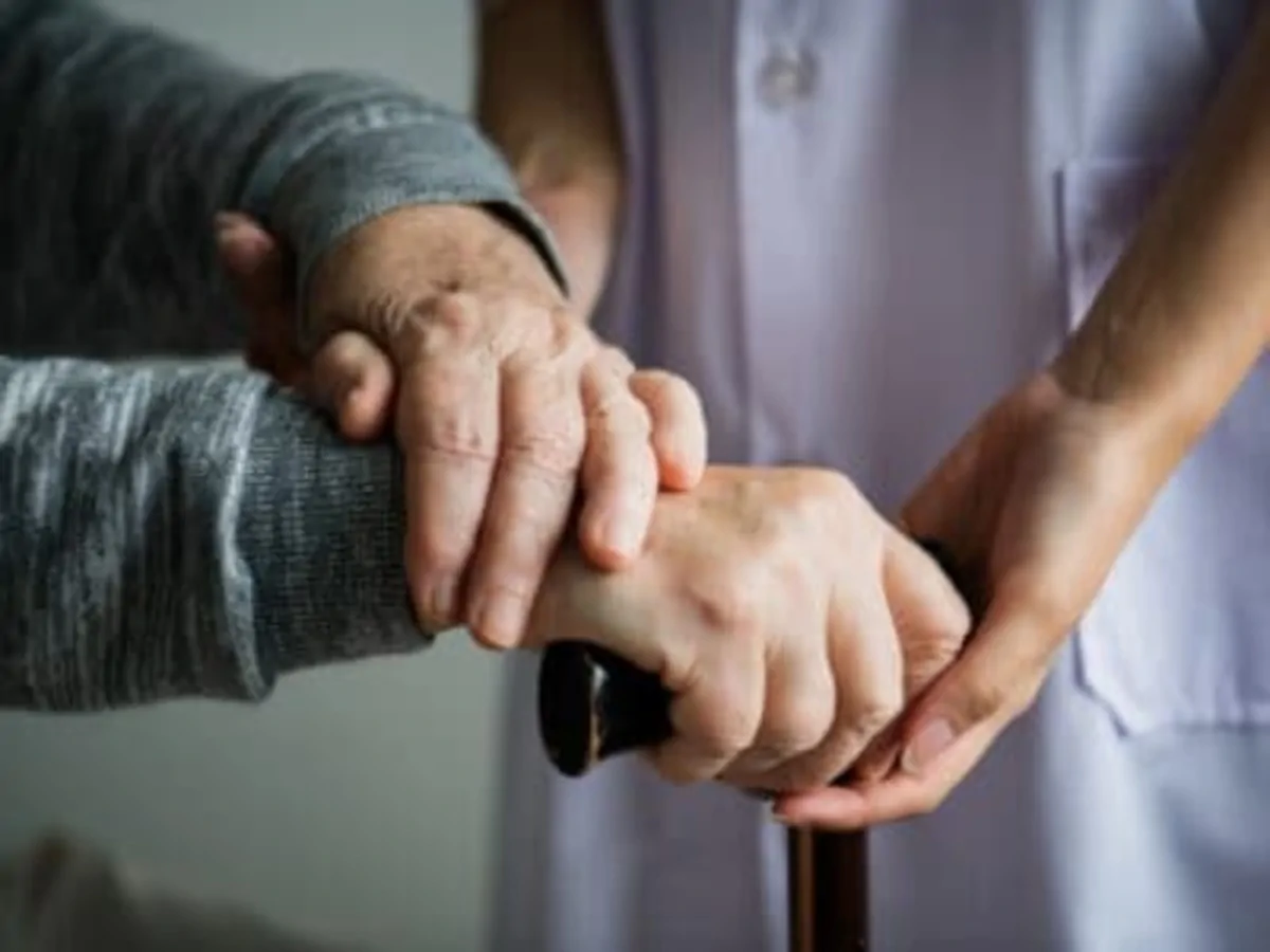 Gros plan sur deux mains entrelacées : une personne âgée avec une canne reçoit le soutien d'un aidant dans un cadre de soins.