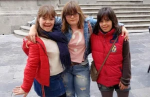 Trois jeunes posent à l'extérieur : l'une porte une veste rouge et une jupe, l'autre porte un t-shirt clair et un jean, la troisième porte une veste rouge.