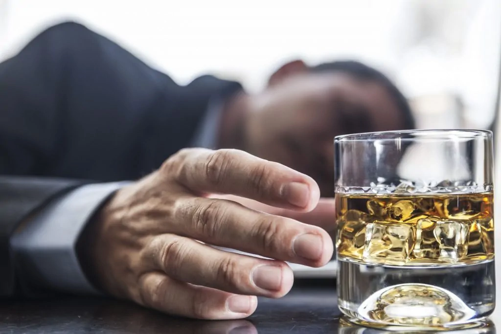 Homme adulte en costume allongé sur une table, regardant un verre de whisky avec des glaçons ; éclairage doux.