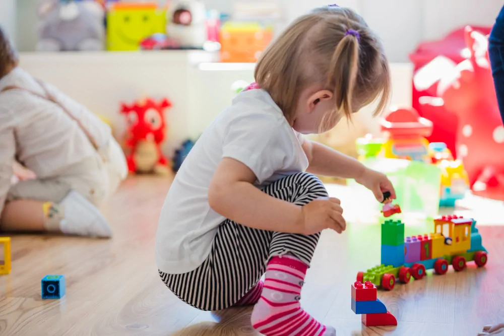 Petite fille assise par terre, construisant avec des blocs de couleurs ; arrière-plan lumineux et présence d'une autre silhouette.
