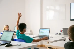 Garçon d'origine mixte portant des lunettes lève la main dans une salle de classe lumineuse, entouré de camarades et d'ordinateurs portables.