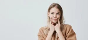 Personne aux cheveux blonds et expression amusée, se tenant la bouche avec les doigts pour forcer un sourire, front blanc.