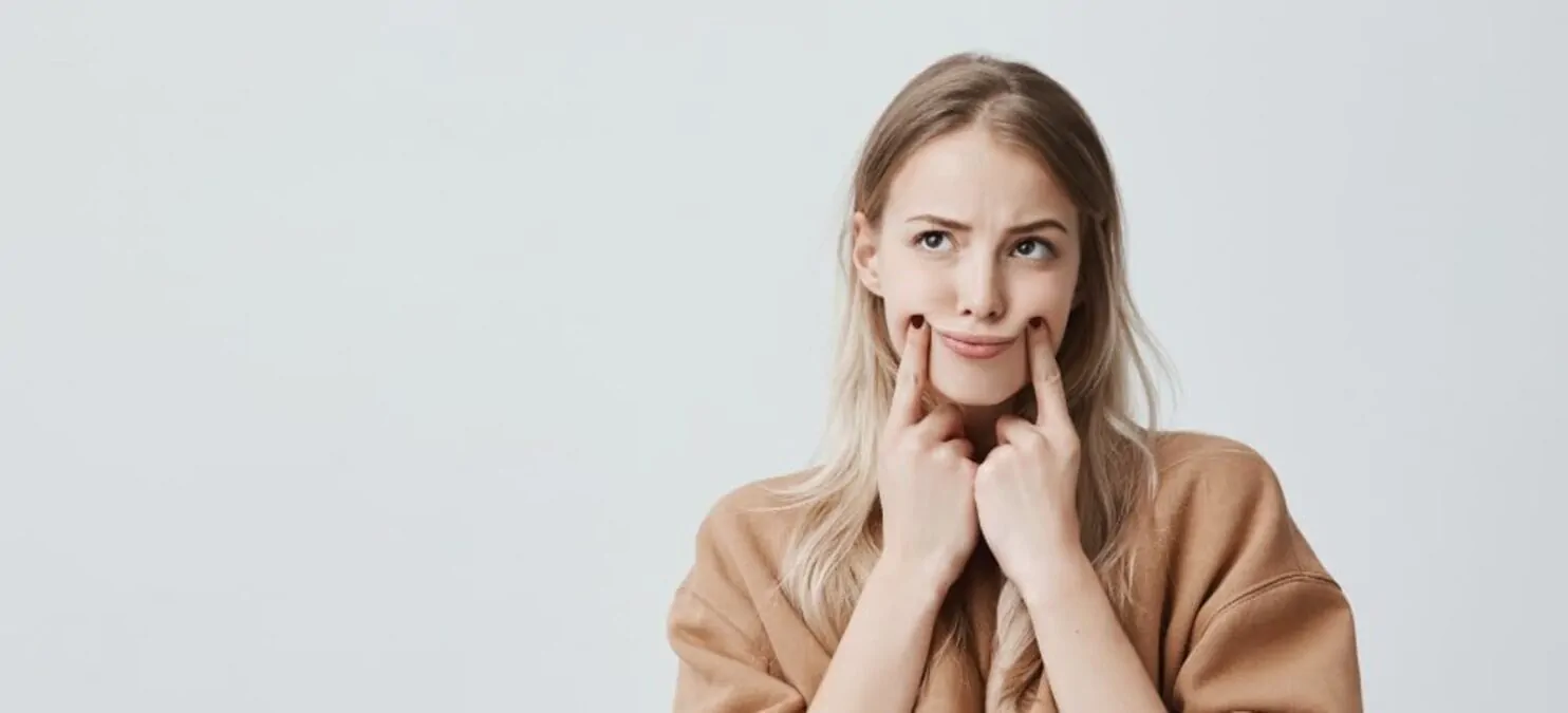 Personne aux cheveux blonds et expression amusée, se tenant la bouche avec les doigts pour forcer un sourire, front blanc.