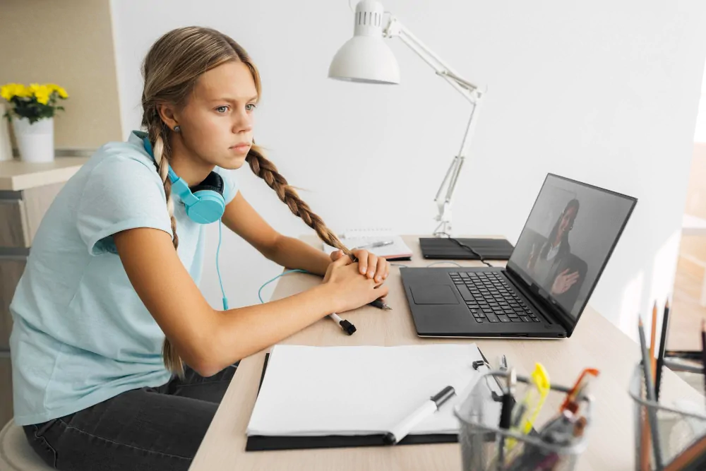 Jeune femme aux tresses portant des écouteurs bleus, devant un ordinateur portable sur un bureau éclairé, avec une lampe, un cahier et une plante.