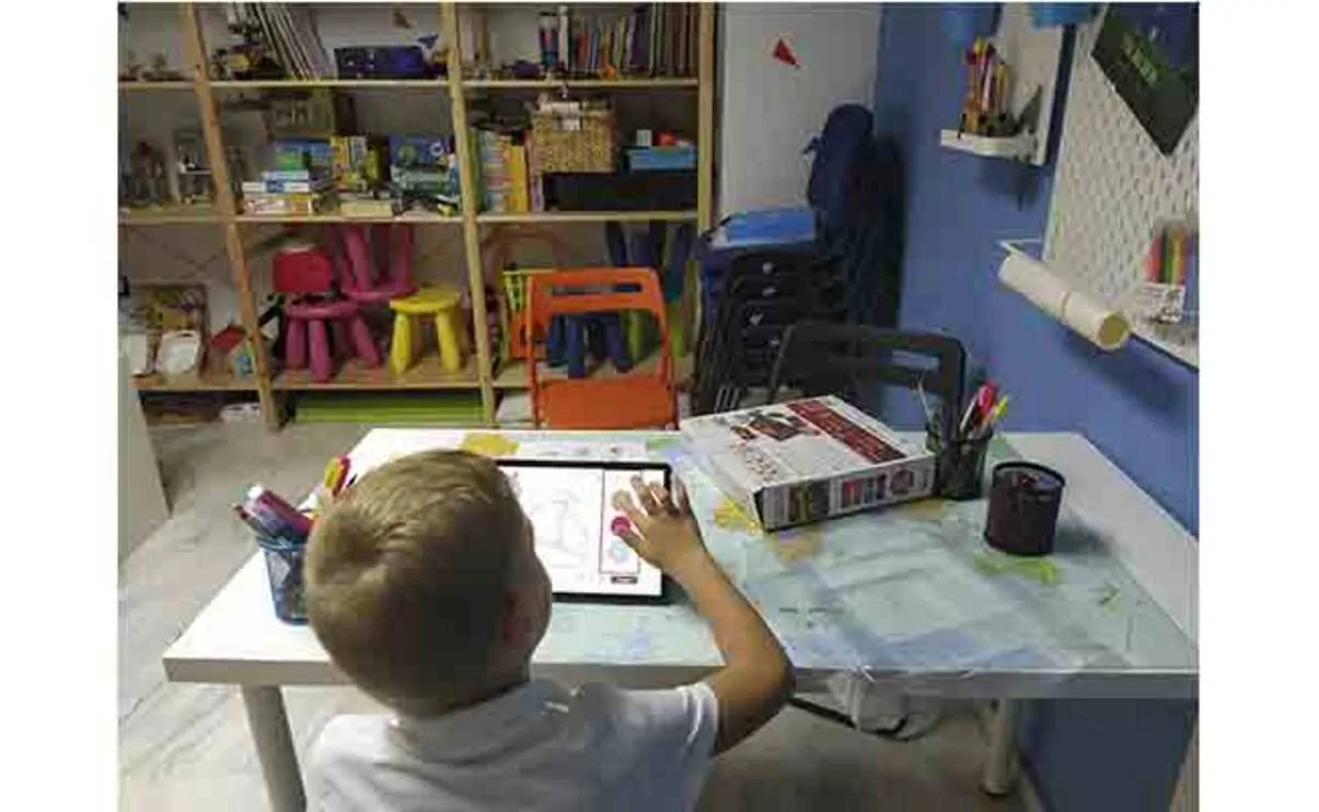 Enfant assis face à une tablette et à une feuille sur une table blanche, dans une salle de classe organisée pour des exercices de stimulation neuropsychologique de la dyslexie.