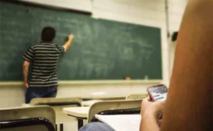 Salle de classe avec un professeur de dos face à un tableau vert; plan rapproché d'un étudiant assis avec son téléphone portable, ambiance de classe.