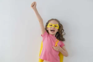 Fille vêtue d'un costume de super-héroïne avec le ruban doré de la Journée mondiale du cancer de l'enfance, regardant l'objectif avec une expression d'espoir.