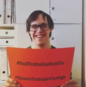 Personne souriante portant des lunettes tient une affiche orange avec '#VullTreballarAmbTu' et '#QuieroTrabajarContigo' dans un bureau.