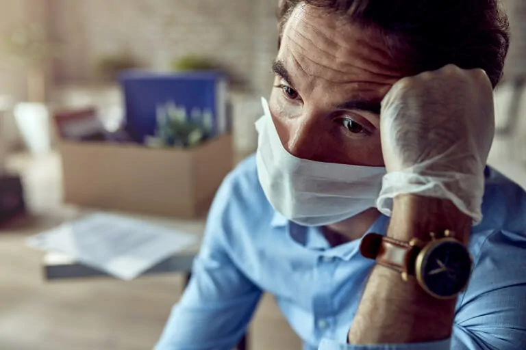 Homme adulte avec masque et gants à l'intérieur, air préoccupé, avec une boîte visible en arrière-plan.