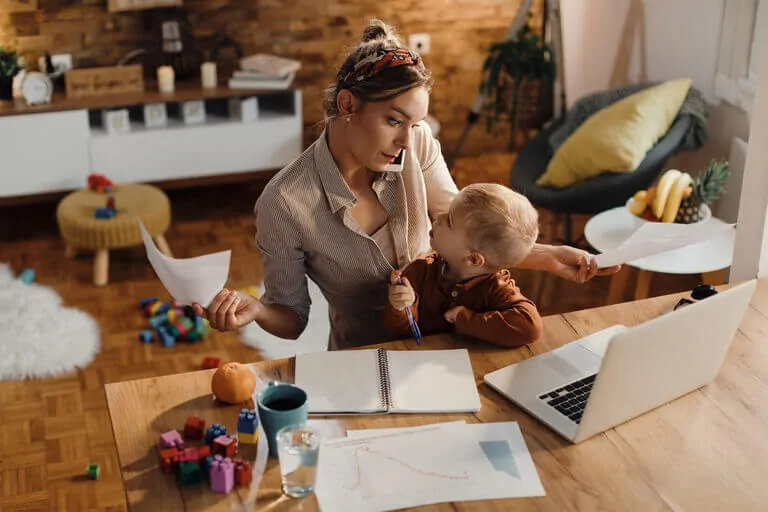 Femme dans un environnement domestique effectuant plusieurs tâches : téléphone, papiers, ordinateur portable ; un enfant observe avec un crayon et des jouets en arrière-plan.