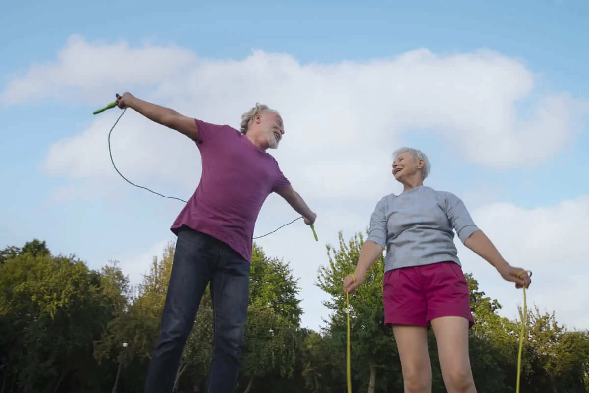 Deux adultes âgés dans un parc faisant de l'exercice ; un homme saute à la corde tandis qu'une femme l'observe, sous un ciel dégagé.