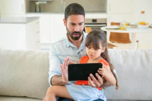 Un homme barbu et une fille assis sur un canapé regardent une tablette, face à une cuisine lumineuse en arrière-plan.