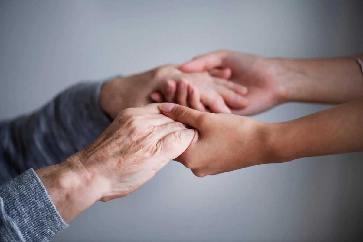 Mains entrelacées au premier plan, une personne âgée et une autre plus jeune, avec un arrière-plan flou.