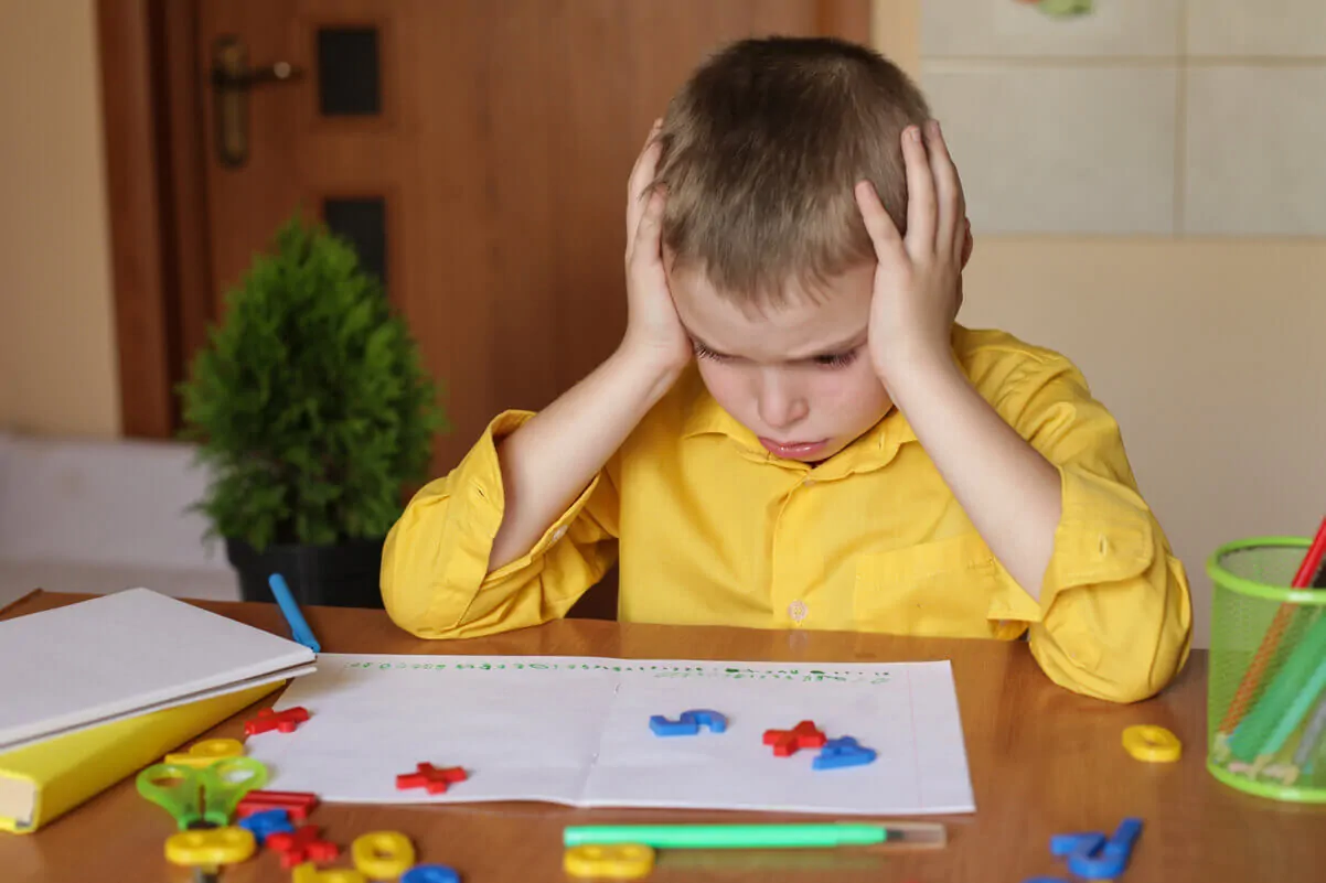 Enfant assis à une table avec une expression de frustration, chemise jaune, cahier ouvert et pièces de couleur éparpillées.