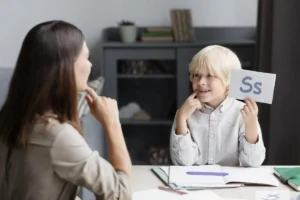 Enfant avec une carte de la lettre S sur une table, supervisé par une professionnelle; intervention éducative avec NeuronUP sur une tablette.