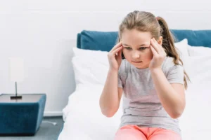 Fille assise sur le lit d'hôpital, regard de malaise et se touche la tête, dans un environnement clinique dédié à la stimulation cognitive.