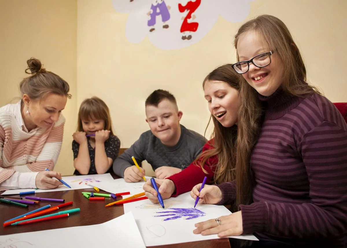 Groupe de trois enfants et deux adultes participant à une activité de bricolage autour d'une table dans un environnement scolaire inclusif.