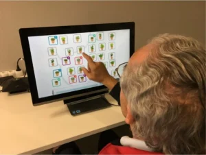 Personne âgée assise devant un écran; à l'écran apparaissent des icônes de pots de fleurs et la personne en désigne un du doigt.