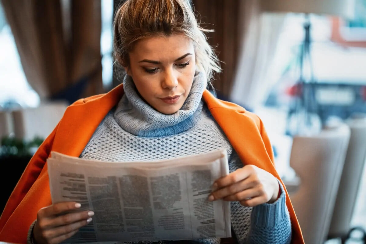 Femme portant un manteau orange assise dans un environnement chaleureux lisant un journal, avec un éclairage doux.