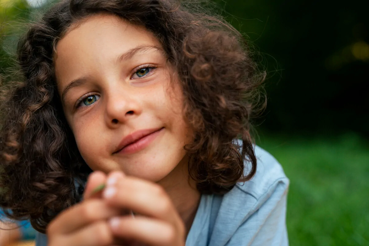 Enfant, garçon ou fille, aux cheveux bruns et bouclés, expression calme et sourire, tenant un objet dans un environnement naturel avec de l'herbe verte.