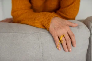 Main reposant sur un canapé clair, avec un pull orange et un ruban jaune au poignet, scène calme pour la SEP.