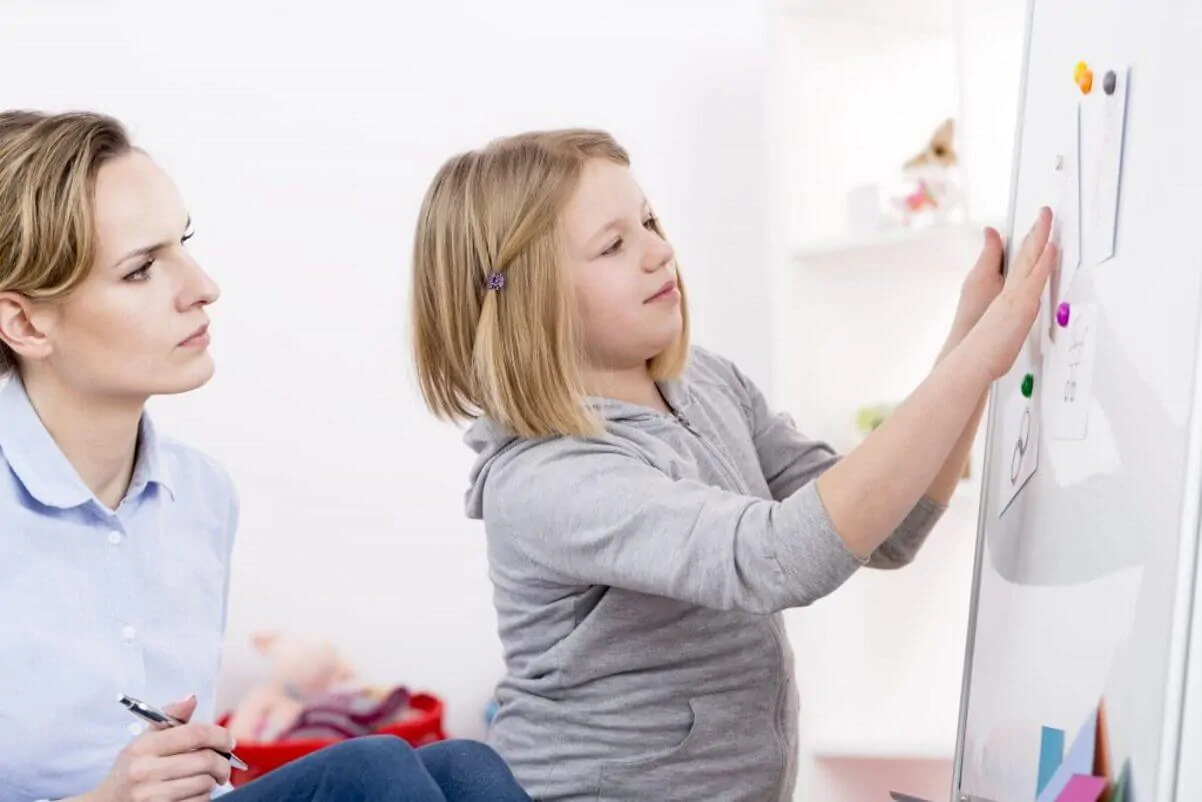 Fille interagit avec un tableau pendant qu'une thérapeute l'observe, dans un environnement éducatif qui illustre la perspective sociale.