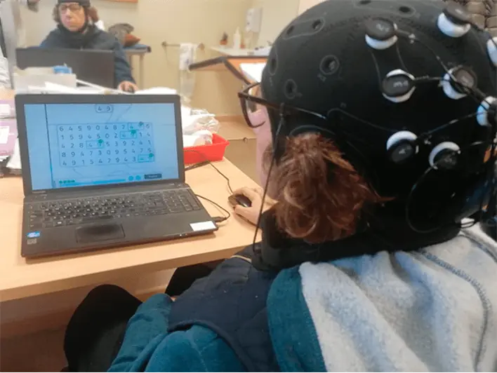 Personne portant une coiffe EEG devant un ordinateur portable ; l'écran affiche une grille de chiffres lors d'une séance de tDCS avec NeuronUP.