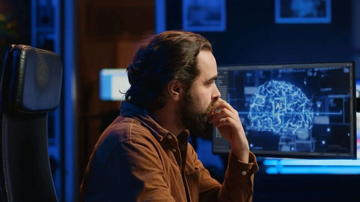 Homme barbu devant un ordinateur, écran bleu avec un graphique du cerveau, ambiance moderne et technologique.