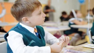 Un enfant en classe, attentif, porte un gilet vert, une chemise blanche et une cravate ; à l'arrière-plan, d'autres élèves.