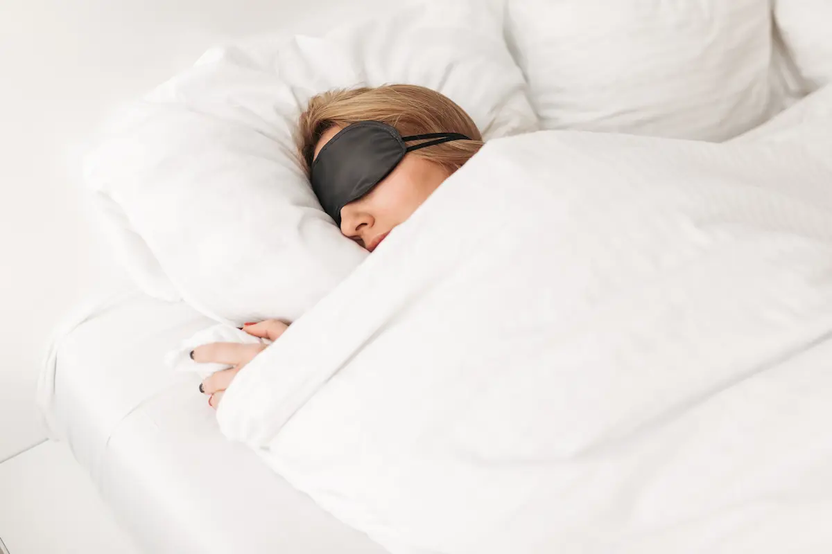 Personne dormant dans un lit avec un masque noir pour les yeux et une couverture blanche, scène sereine illustrant le sommeil et la vigilance cognitive.
