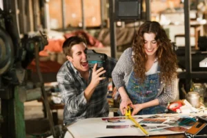 Deux personnes travaillant dans un atelier créatif : l'une filme avec une caméra et l'autre découpe du papier, toutes deux souriantes.