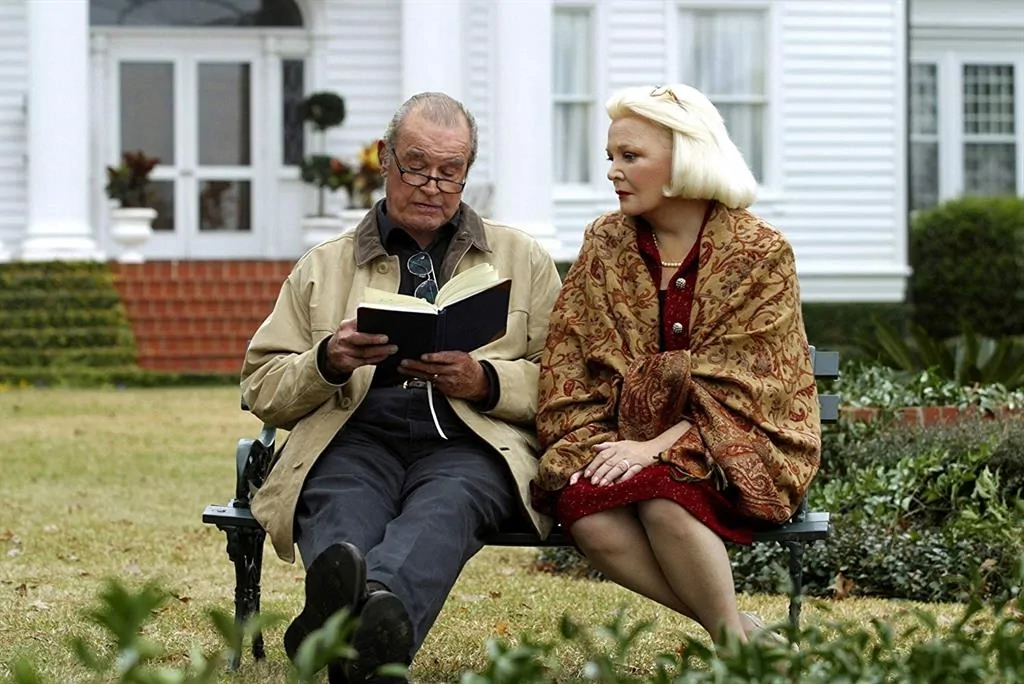 Deux personnes assises sur un banc de jardin ; l'homme lit un livre et la femme l'observe, avec une maison blanche en arrière-plan.