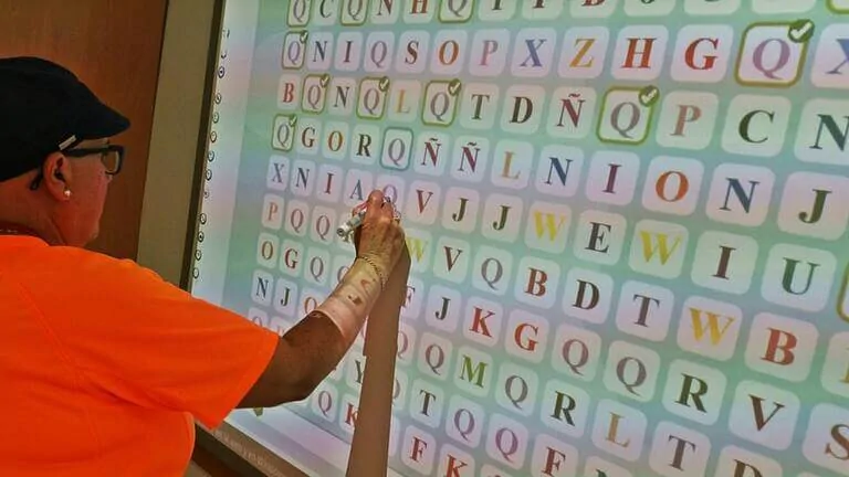 Persona anziana in ambiente clinico di riabilitazione che interagisce con uno schermo di lettere colorate, indicando per stimolazione cognitiva.