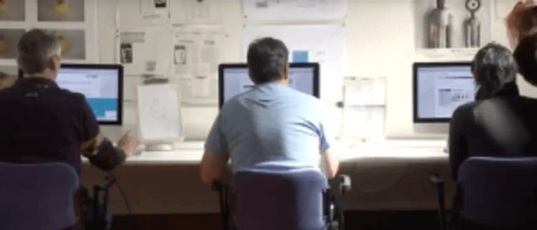 Gruppo di persone sedute davanti a computer in un ambiente di lavoro, che osservano schermi con contenuti digitali e grafici.