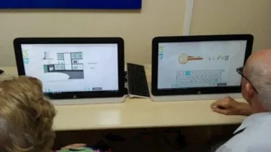 Due computer su tavolo educativo; gli schermi mostrano un progetto di cucina e un tasto, con due utenti di fronte a essi.